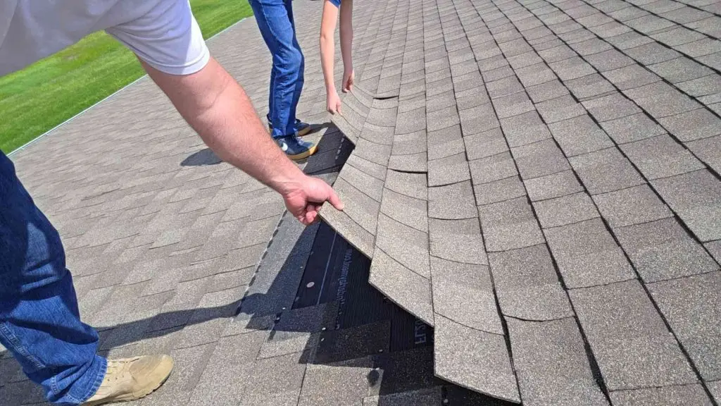 Two people inspecting damaged gray asphalt roof shingles.