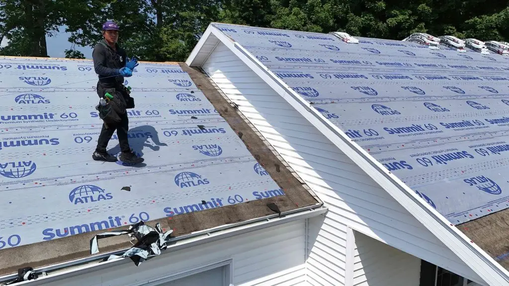 Roofer on a roof, installing roofing underlayment. Gray underlayment, house with white trim, sunny day.