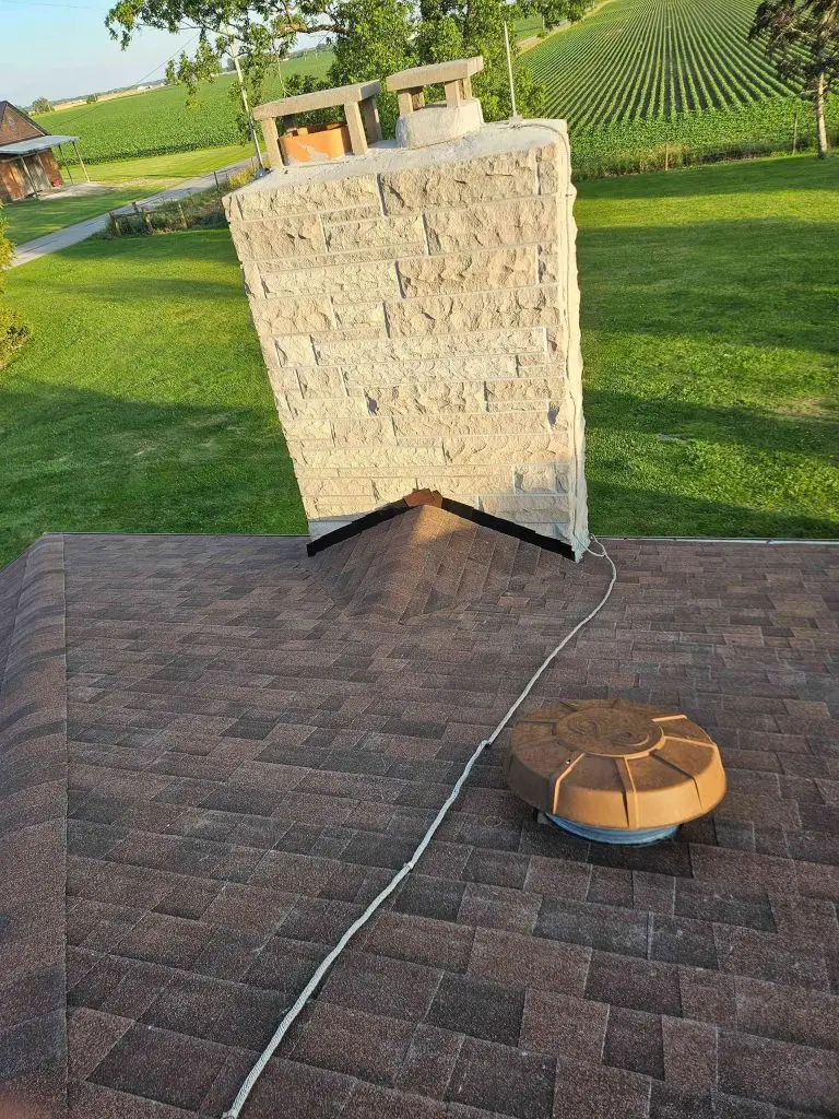 Leaning chimney on a brown shingle roof, cable to a round vent. Green field in background.