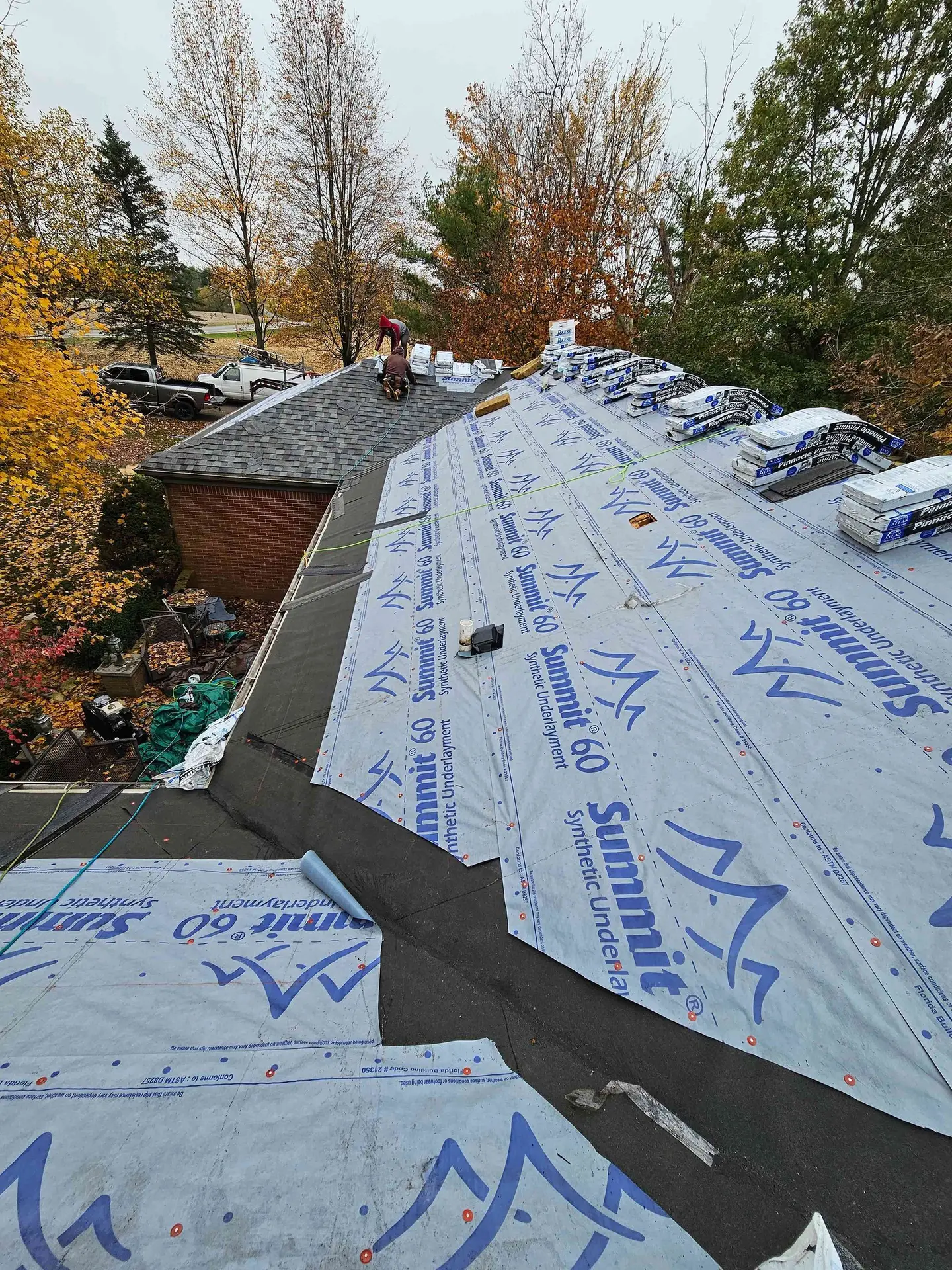 Roofers installing shingles on a house roof, surrounded by trees with fall foliage.