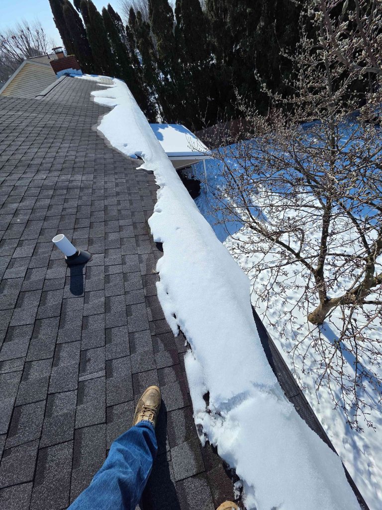 Snow-covered roof edge; a person's legs and feet are visible. Blue jeans, brown boots. Sunny, winter day.