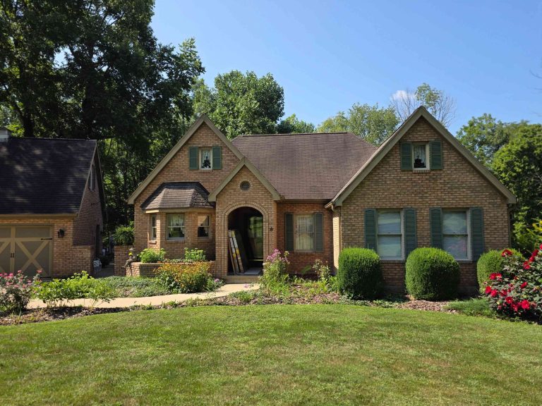 Brick house with green shutters and brown roof; flowers in the front yard.
