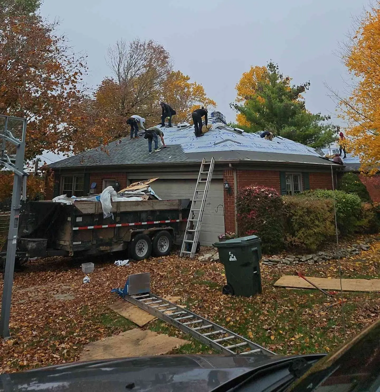 Roofing workers on a house roof removing shingles with a dumpster trailer and ladders. Autumn leaves surround.