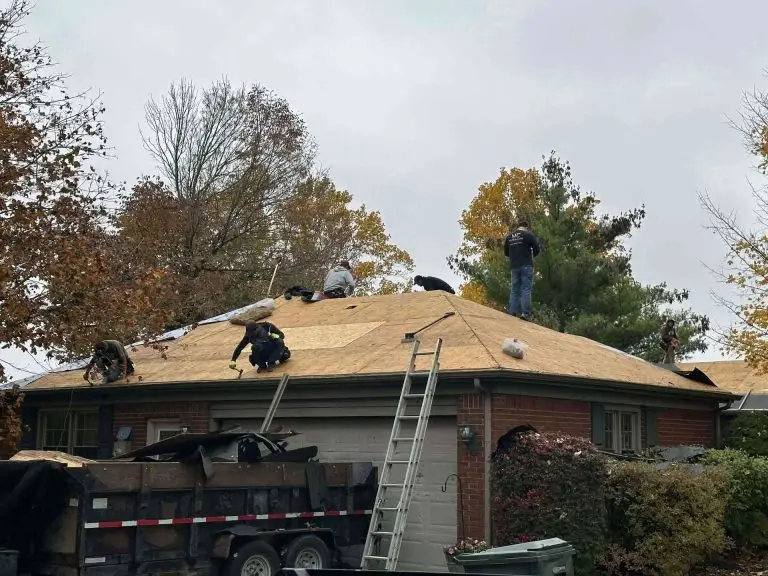 Roofing crew working on a house roof. A ladder leans against the roof. Trailer with debris in front.