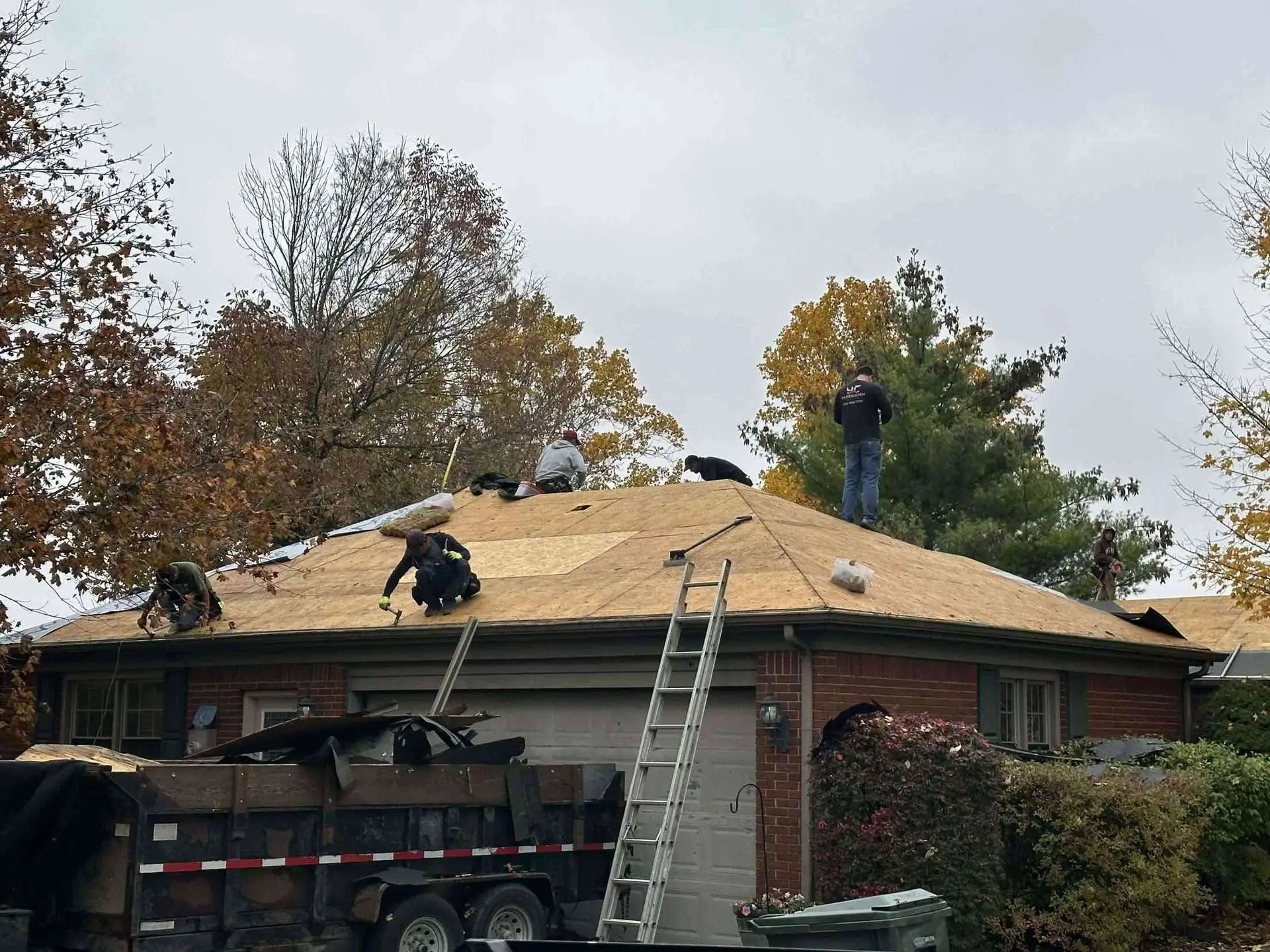 Roofing crew working on a house roof. A ladder leans against the roof. Trailer with debris in front.