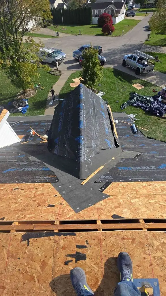 Roofer on a partially shingled roof, looking down. Roofing materials are visible. Suburban neighborhood in the background.